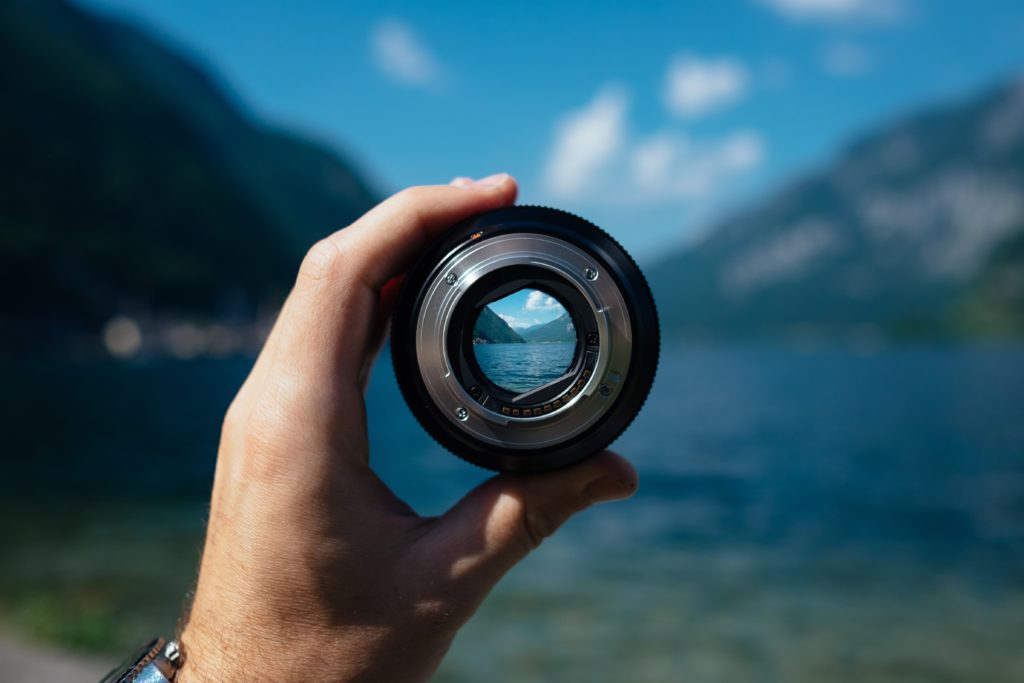 Image of a man's hand holding a camera lens in front of a landscape, through which the scene can be seen in focus