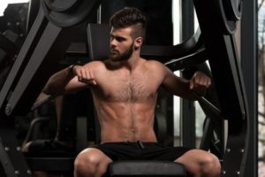 Photo of a shirtless, bearded man sitting at a chest press machine in a dark gym.