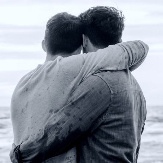 Photo of two men from the back, embracing and staring out to sea