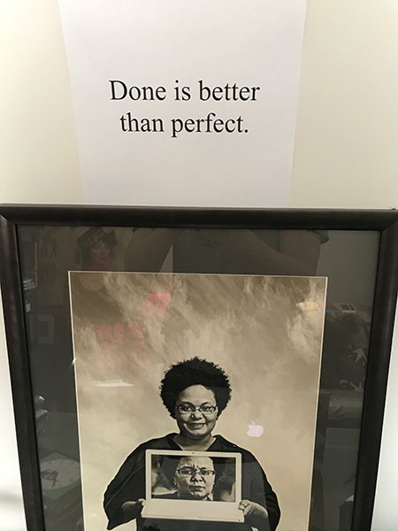 The phrase "Done is better than perfect" taped to the wall above a photo of my friend Pamela, a Black woman smiling and holding a laptop that displays her face giving the viewer side-eye.