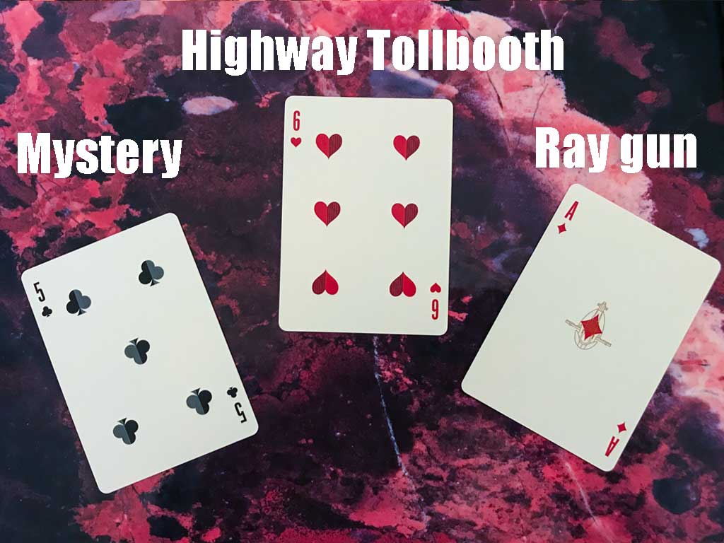 Three playing cards—a five of clubs, a six of hearts, and an ace of diamonds—laid against a red marbled background form the basis of a writing prompt: a mystery, set in a highway tollbooth, featuring a ray gun.