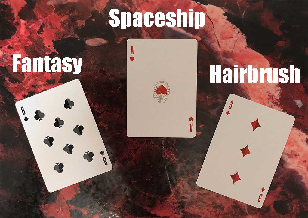 Photo of three playing cards on a red marbled color background. The cards are the eight of clubs, over which is the word "fantasy"; the ace of hearts, over which is the word "spaceship"; and the three of diamonds, over which is the word "hairbrush." 